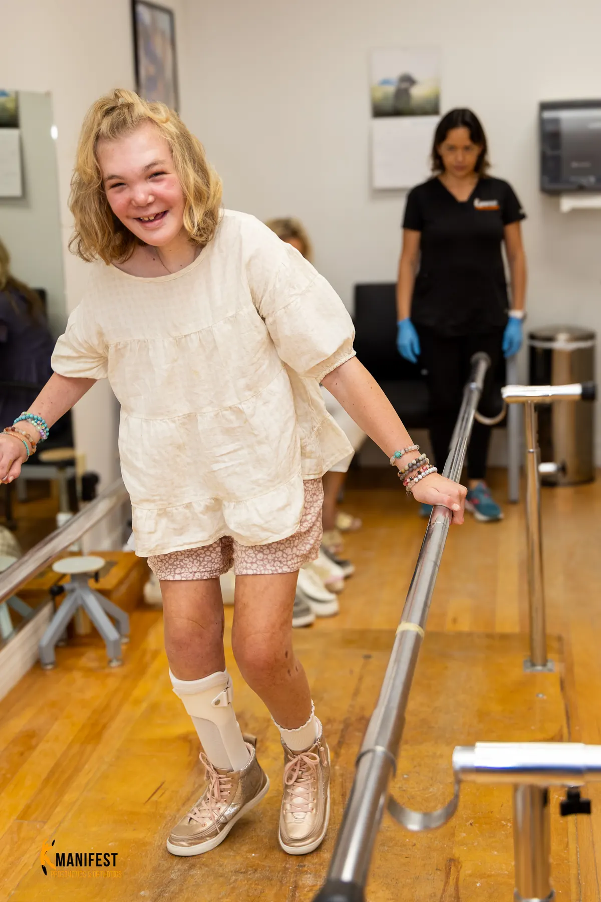 A young Manifest patient walking with a custom ankle-foot orthosis, smiling at the camera with her clinician supporting in the background.